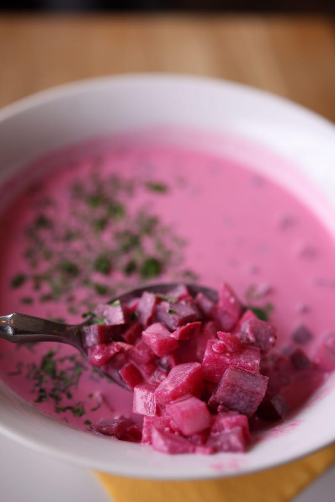 Close-up of a bowl of vibrant pink beetroot soup with parsley garnish and a spoonful of diced beets.