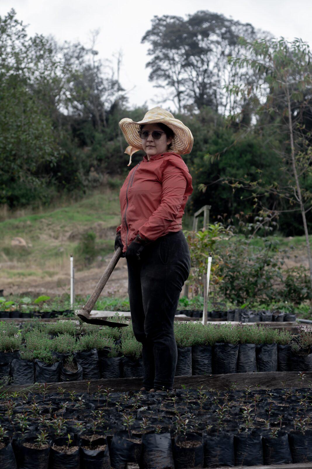 Woman in sunhat and sunglasses working on a lush farm, carrying a hoe among plants.