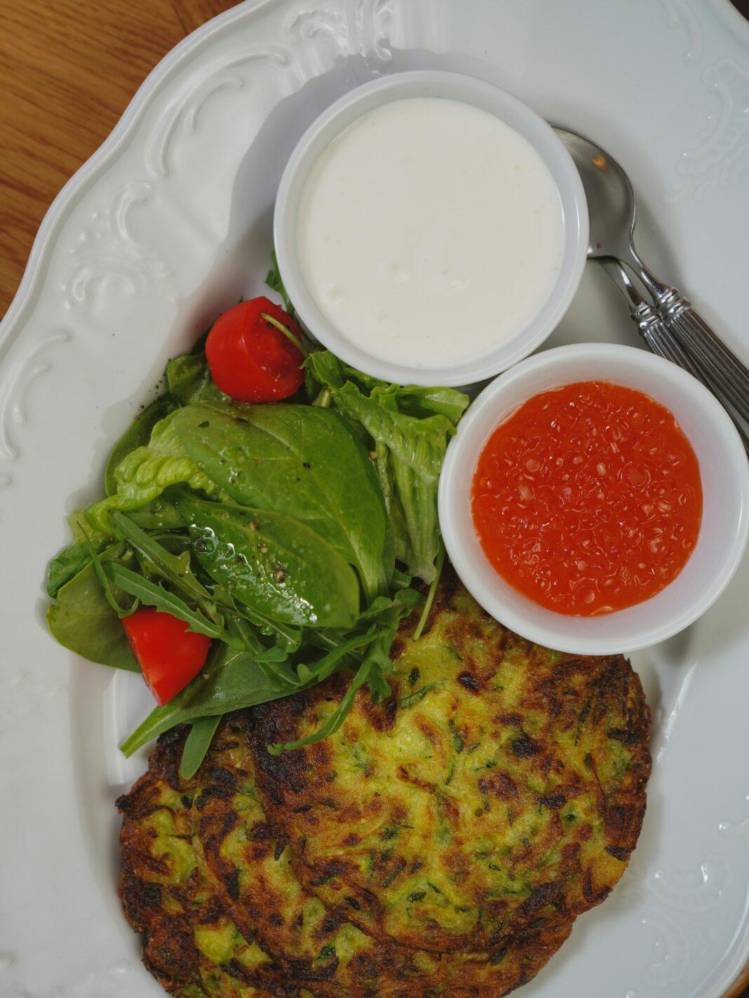 A tasty zucchini fritter served with fresh salad and two distinct sauces on a white plate.