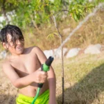 Arrosage des arbres fruitiers en été : 10 à 20 litres pour un jeune arbre, pas pour tous A cheerful child enjoys a sunny afternoon watering plants with a hose in a garden.