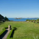 Plantes de bord de mer : 3 étapes avant de planter, le vent tranche A peaceful coastal garden with lush greenery overlooking a calm sea under a clear blue sky.