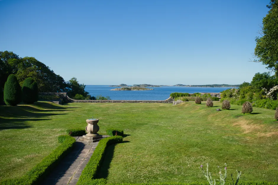 A peaceful coastal garden with lush greenery overlooking a calm sea under a clear blue sky.