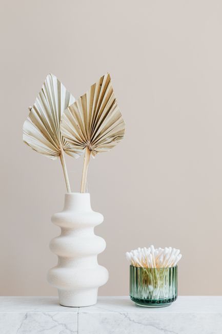 Composition of white ceramic vase with artificial leaves and green glass reusable container for cotton buds placed on white marble shelf against beige wall