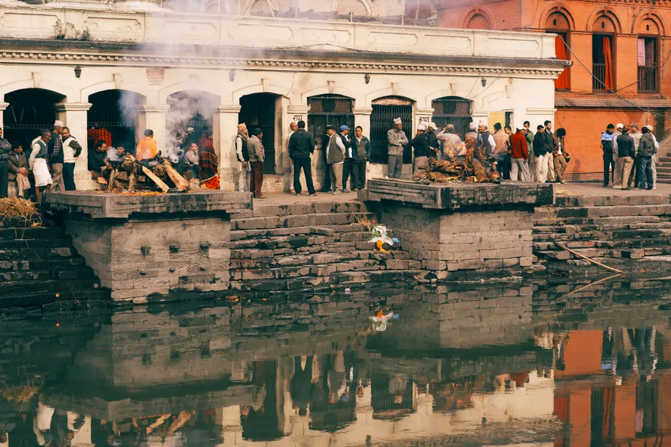 Outdoor scene of a traditional cremation ceremony near a river with a crowd of people.