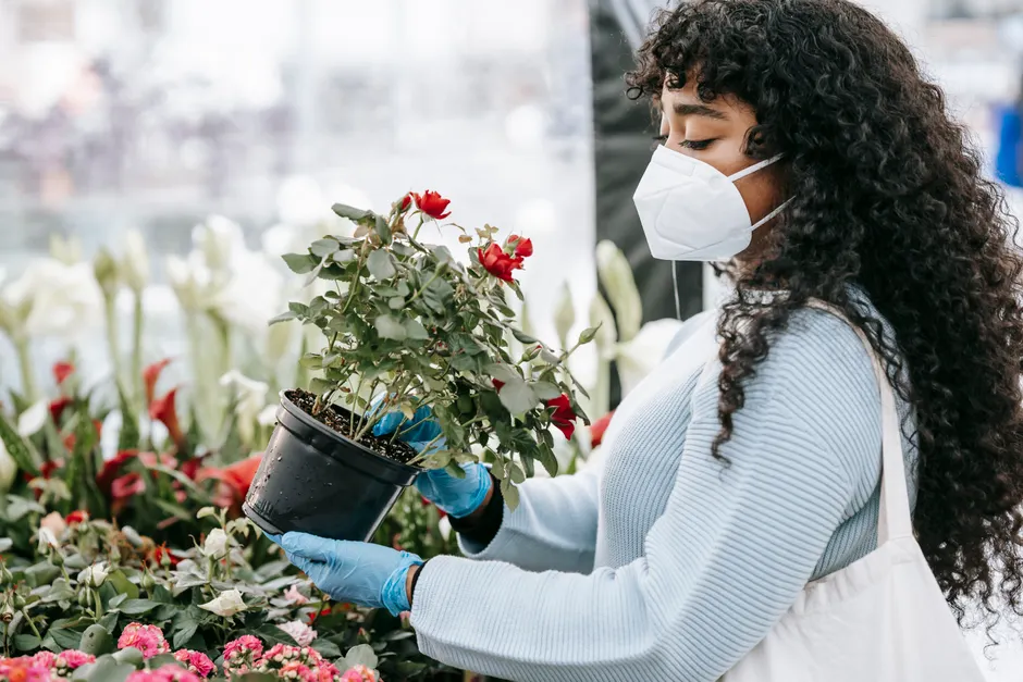 Side view crop African American female in respirator and gloves choosing blooming red potted flowers in street market