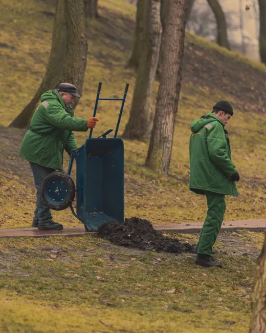 Two gardeners in green uniforms using a wheelbarrow in a park setting.
