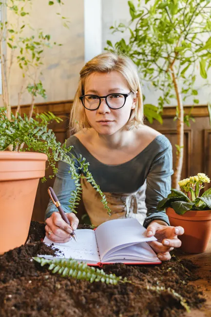 A young woman takes gardening notes surrounded by plants indoors, blending nature with study.