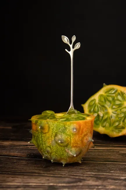 Vibrant close-up of a horned melon (kiwano) with a silver spoon on a wooden surface.