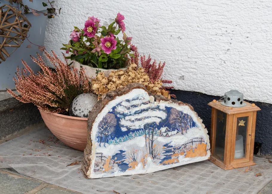 A cozy outdoor corner with potted plants and a decorative winter scene on stone, alongside a rustic lantern.