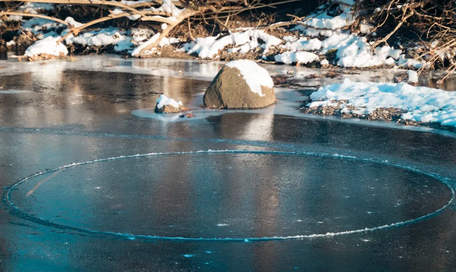 A frozen winter pond with a natural ice circle and snow-covered rocks.
