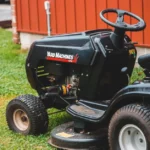 Tracteur tondeuse : enfants hors zone, sans exception Close-up of a black riding lawn mower parked on a lawn next to a red building.