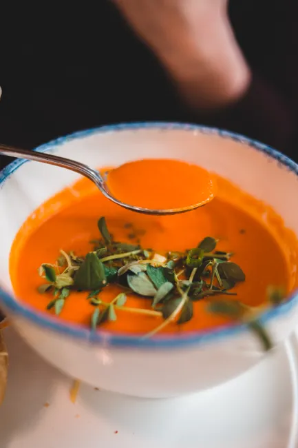From above of crop unrecognizable person dining with appetizing vegetable tomato soup decorated with fresh watercress leaves in ceramic bowl in cafe