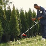 Débroussailleuse en 2026 : le terrain tranche, pas la puissance Man using grass cutter to trim trees in a sunny outdoor garden.